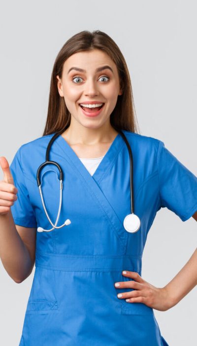 Medical workers, healthcare, covid-19 and vaccination concept. Surprised excited female nurse or doctor in blue scrubs with stethoscope, show thumbs-up in approval, like idea, smiling upbeat.